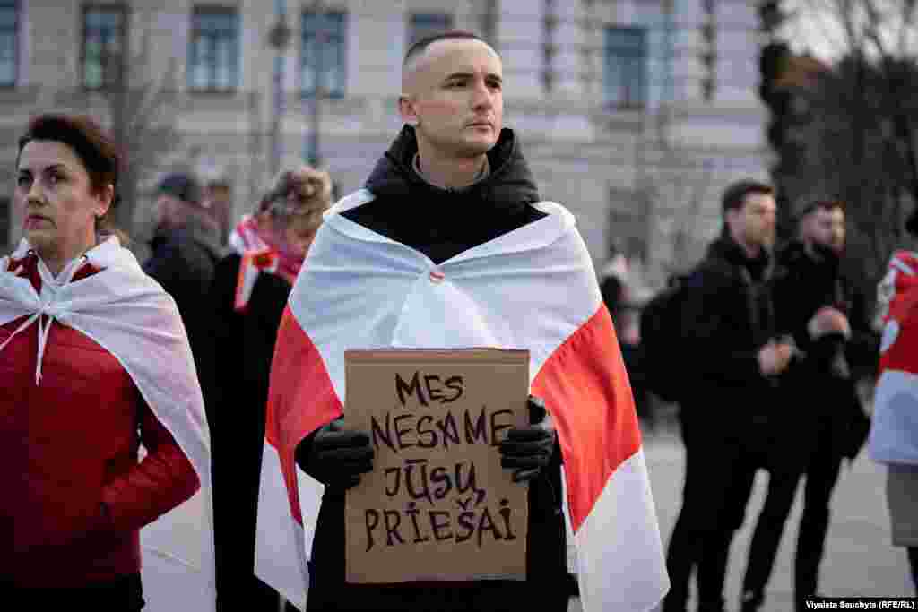 A man draped in the white-red-white flag holds a sign that reads, "We are not your enemies." The widespread protests were met with a harsh crackdown by the government, resulting in the arrests of opposition figures and activists.
